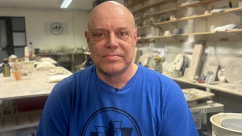 Alan Dunnighan sits facing the camera. He is bald and wears a blue T-shirt with a design in a black circle which can be partially seen in the shot. Behind him  is a pottery studio which consists of a large table with pieces of equipment and shelves containing pieces of pottery.