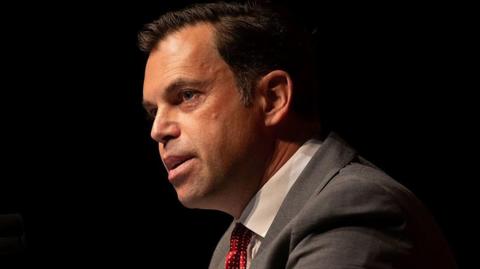 Ken Skates in front of a black backdrop. He is wearing a grey suit and a red tie