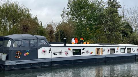 A barge sits on a canal with trees behind it. It is black and white, but is decorated along it's length with Halloween decorations stuck to it's side including spiders, cobwebs and bats.