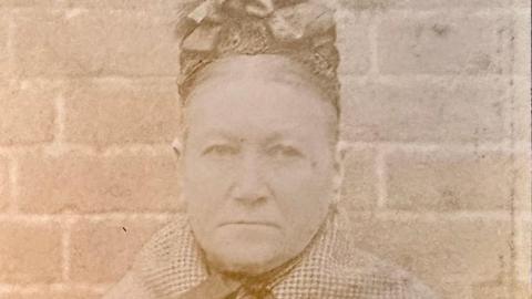 A sepia close-up photograph of a stern faced heavily built woman in her fifties with her hair parted in the middle and mostly hidden by a hat made out of lace with bows on the top. She is wearing a checked shawl and standing in front of a brick wall.
