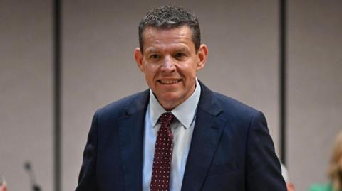 Rhun ap Iorwerth, a man with short brown hair in a navy suit, speaks while standing at a lectern.