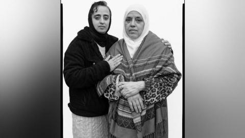 A black and white portrait of two women. One has very short dark hair as well as a black head scarf. She is wearing a dark jacket, and white skirt. She has her arm around another woman who has a white head scarf on, and a patterned shawl