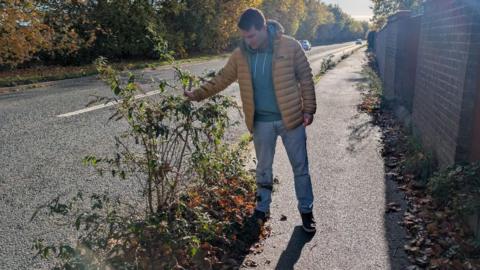 Councillor Nathan Ley on Twelve Acre Drive, Abingdon, holding up some shoulder-length weeds sprouting from a pavement next to a main road.