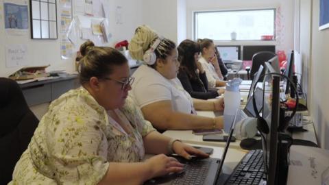Five women sit on a long desk facing computer screens.