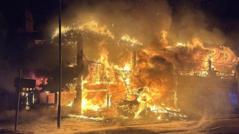 A fire is earing through a two storey building and has burnt down the walls, just leaving the bones and foundations of the site. It remains engulfed in flames. The sky is dark making the fire appear even brighter against the night sky.