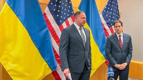 Andriy Yermak and Marco Rubio stand in front of Ukrainian and US flags in a conference room. Yermak is wearing a grey tie, and Rubio a red one.