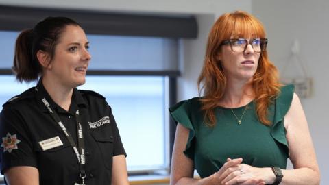 A woman with dark hair tied in a ponytail is wearing a black t-shirt and lanyard. her t-shirt says "fire control". next to her in angela rayner, who has bright ginger hair and is wearing a green dress