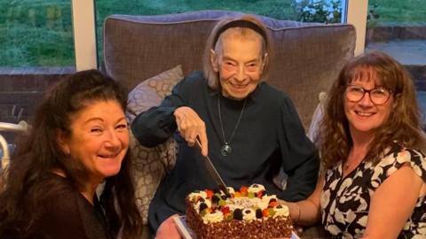 An elderly woman sat on a sofa cutting into her birthday cake that has lots of fruit on it.  She has short hair that has been pushed back with a headband and she is wearing a dark black jumper and a long necklace. She is smiling at the camera. On the right another woman is holding the cake while kneeling next to the elderly woman. She is wearing a sleeveless black top with white flowers and she is smiling at the camera while wearing black frame glasses. She has curly brown hair. Another woman is kneeling on the other side and has black curly hair and is also smiling at the camera.