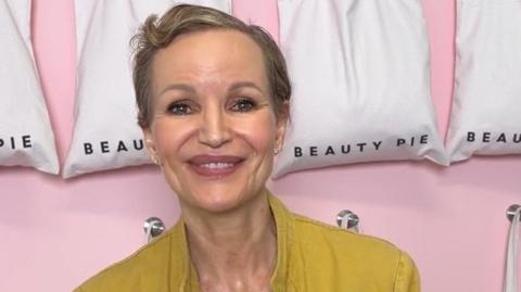 Marcia Kilgore with cropped short brown hair smiling in front of a row of white bag with the Beauty Pie logo on thems at the camera with a blurred background wearing a nude coloured round neck top