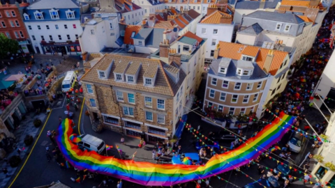 A birds eye view of a street. There is a large rainbow flag being carried through the street with many small rainbow flags and bunting. There are crowds. There are buildings around. 