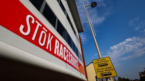 Outside of the STōK Racecourse. A red banner is across the building which reads 'STōK Racecourse' in capital letters. The font is bold and white. A lamp post can be seen next to the building which has a yellow and black coach drop-off sign on it.