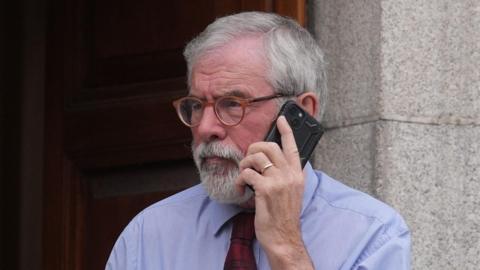 Former Sinn Fein president Gerry Adams holding a mobile phone to his ear outside the High Court in Dublin.  He has short, grey hair and a grey beard.  He is wearing brown-rimmed glasses, a blue shirt and a red patterned tie. 