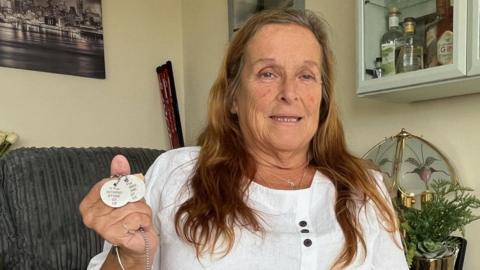Angie Stone, a women in her 60s, with long brown hair, is holding two identification disks, on a chain, stamped with Gregg Stone's Army number, name and religion.