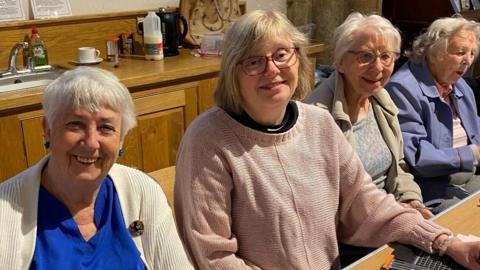 Four women sat next to each other smiling. The woman on the far left has a bright blue top and blue earrings, next to her is a woman wearing a pink knitted jumper and glasses. Next to that woman is a women wearing a blue top and beige fleece with glasses. On the far right is a woman wearing a purple jacket. They are sat inside with a sink behind them and milk, coffee and a kettle.