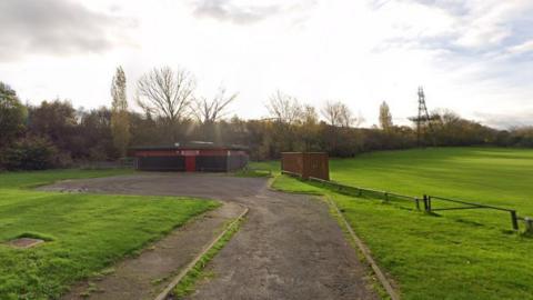 A grass playing field, with a car park in the foreground. Also visible is a single-storey clubhouse painted red.