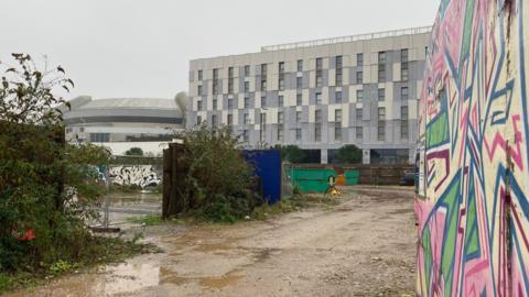 A building site on a wet day in Plymouth with fences around parts of the site and a wall with pink, green and blue graffiti drawn on it and a block of flats in the background.