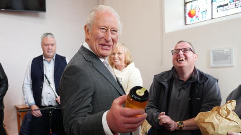 The King wears a dark-grey suit, white shirt and grey tie. He holds a clear jar of honey in his left hand. Her laughs as three people stand behind him. They are inside a building with white walls. A TV is over the King's right shoulder and a stain-glass window is over his left.