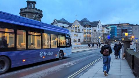 A purple First Bus travels across the Victorian iron Lendal Bridge while pedestrians walk along the pavement past the medieval Lendal Tower under an overcast winter sky.