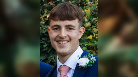 Ethan Powell wearing a navy suit and pink tie. He is smiling at the camera. 