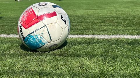 A close up of a football on a pitch. The ball is white with red and blue marks and Joma branding on it. It's on a green football pitch with a white line painted along it. 