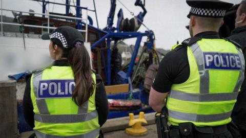 Police stand next to a blue trawler vessel moored in a harbour