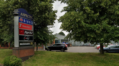 A view of the leisure park from the road with a sign listing a McDonald's, bingo hall, cinema, gym and pizza restaurant. There are cars in a car park behind the sign with the cinema in the background.