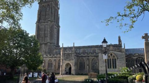 St John's Church on a summer's day. Several people can be seen standing on the pavement besides the black railings outside. There are trees to the left and bunting to the right.