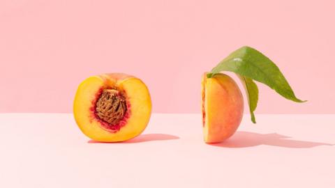 Peach cut in half, the left side is facing the front with the stone in it, the other half is on it's side with the leaf coming out of the top. The peach sits on a pale pink background. 