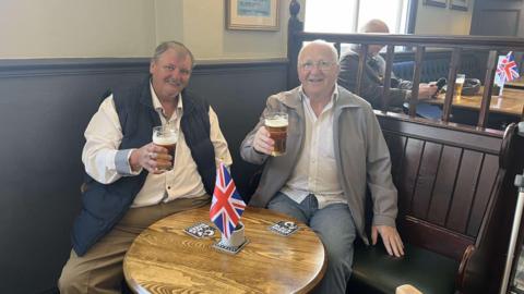 Two men sit at a pub table with union flags around them. They are smiling as they each hold a partly drink pint of beer.