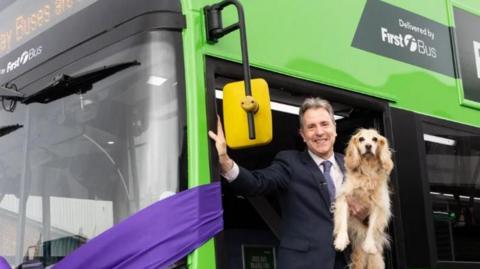 Dan Norris holding his dog while stepping off a green bus with a purple ribbon wrapped around the front.