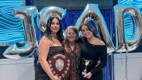 Jeannine, Zoe and Josie all smile at the camera at an awards event as Zoe and Josie hold awards and trophies with Jeannine in the middle. They're arms and around each other. Behind them is are balloons that spell out JSAD.