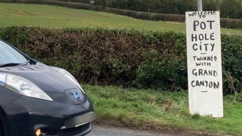 "Welcome to Pothole City - twinned with Grand Canyon" sign on a grass verge.  A car is passing by on the road.