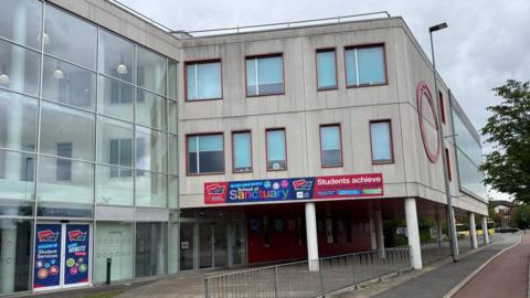 A glass-fronted school building extends towards some tower blocks on a grey day, as seen from across a roadway. 