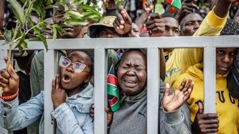 A crowd of people look through grey railings at the main stadium in Kisumu. A woman on the left holds a branch and is chanting. The woman next to her holds a Kenyan flag and has her eyes closed as she weeps - 18 October 2025.