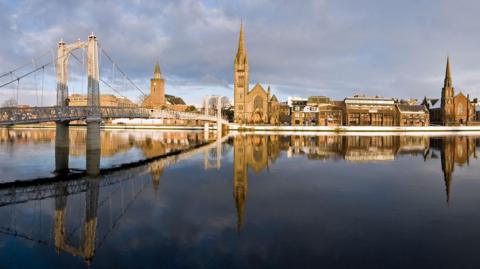 Historic buildings on Inverness' riverside, including churches, reflected in the mirror-like surface of the River Ness on a bright but cloudy day.