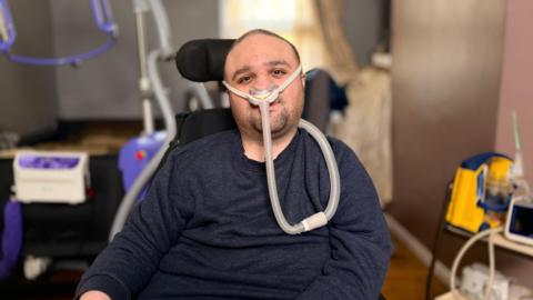 Man in his thirties with short dark hair and stubble, wearing a dark blue jumper looks at camera. He is sitting in a powered wheelchair with a ventilation tube attached to his nose. In the background is medical and hoisting equipment.