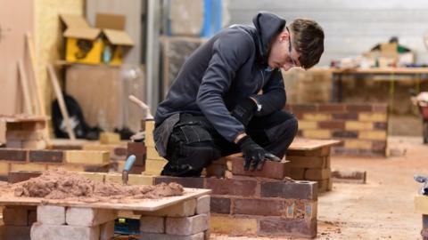 A young man is bent over doing some bricklaying - he is wearing protective glasses and has gloves on. He is holding one brick, which he is pressing onto a row of brickwork.