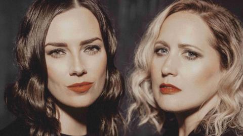 Marissa Keltie, who has dark hair, and Caroline Gilmour, who has blonde hair, look directly into the camera.