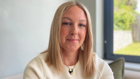 A woman with long blonde hair and brown eyelashes smiles to the camera. She wears a cream buttoned cardigan with a gold chain around her neck. Over her left shoulder is a window in which a green-grass garden can be seen. 