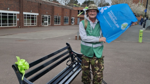 A man wearing a green high-vis jacket, a grey hoodie and camouflage trousers and a hat, holding a blue flag reading "Walking for Mitchs Law" stands next to a black bench with a green bow on it. There is a building behind him