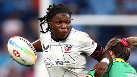 Cheta Emba wears a white USA women's rugby jersey and black stretchy hairband during a match. She is holding a rugby ball with brightly-coloured logos on it in her right hand while trying to fend off the tackle of an opponent in green with her other arm. Emba has a look of intense concentration on her face as she braces for the impact