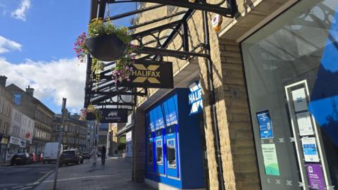 Halifax bank branch on a town-centre street, with bright blue ATM machines and signage set into a sandstone building beneath decorative black metal canopies. Hanging flower baskets line the pavement, while cars, a white van and a few pedestrians are visible along the street. Historic stone buildings extend into the background under a clear blue sky with scattered clouds.