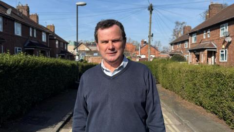 City of York Council's Labour deputy leader Pete Kilbane. He is looking straight at the camera, has receding brown hair, and is wearing a blue jumper and an open neck shirt. He is standing on a hedge-lined street in York. The sun is out and blue sky is in the background.