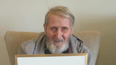 Man with grey beard and wearing grey top looks at camera