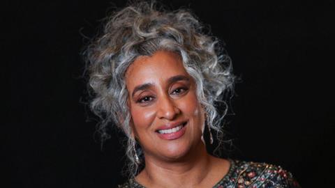 A woman with silver hair and a multicoloured beaded dress smiles.