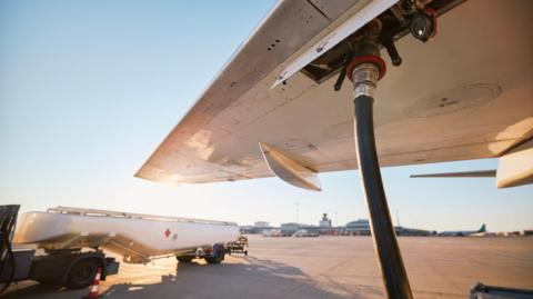 A close up of a plane wing with a jet fuel hose plugged in, and a fuel lorry in the background.