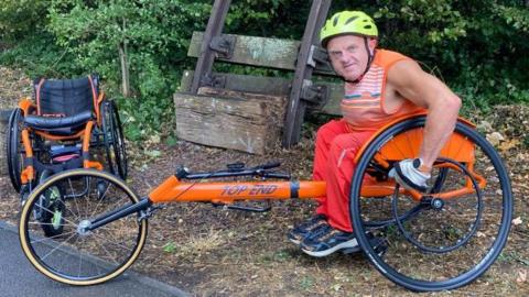A picture of a man in an orange racing wheelchair, wearing orange and a yellow helmet. He is pictured at the side of a path in a park with a second smaller orange wheelchair pictured in the background.