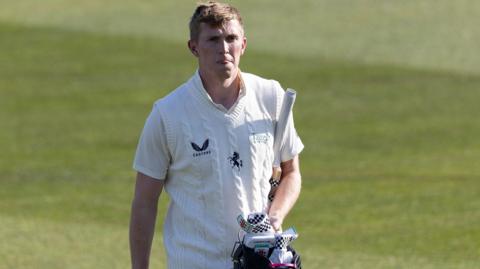 Zak Crawley walks off the cricket ground, dressed in a white short-sleeved top and white knitted vest, carrying a blue helmet and white and black gloves