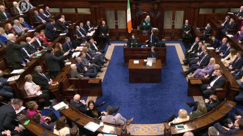 Wide shot of the Dáil with TDs in benches and an Irish flag and protests as the speaker stands up n her chair. 