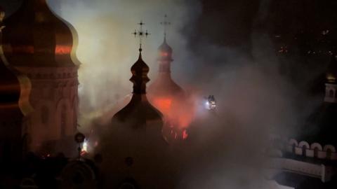 At night, fire glows on the cathedral roof as firefighters on a long ladder spray water on the site.